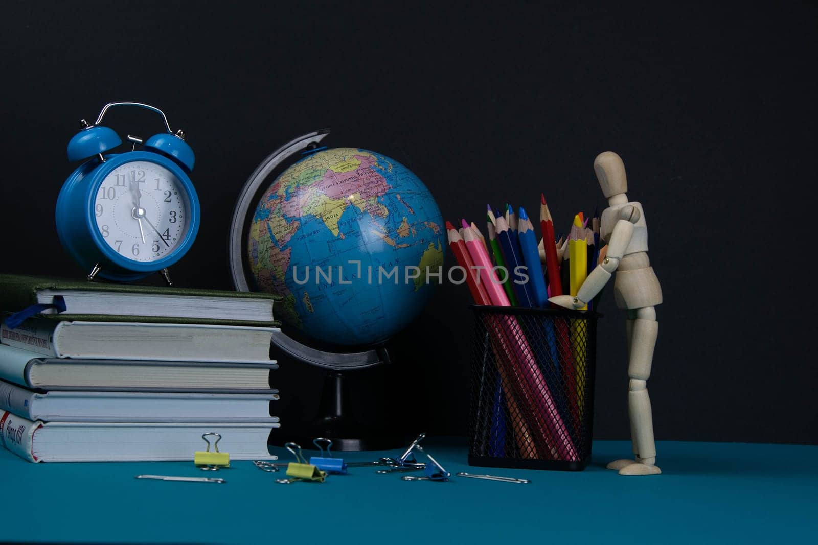 The globe and the clock stand on a stack of books, next to a glass of pencils and the pyramid is folded with wooden cubes and the figure of a wooden man hugs pencils.