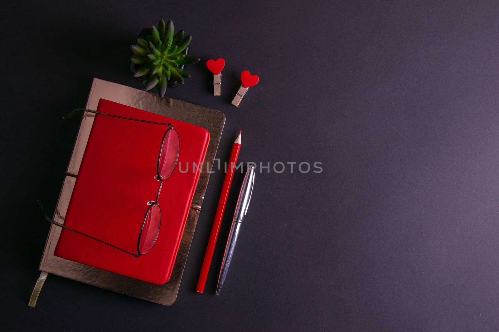 The red notebook stands on a gold notebook which stands on a black background, next to the notebook are a silver pen, a red pencil, two clothespins in the shape of a heart and glasses with a green plant.