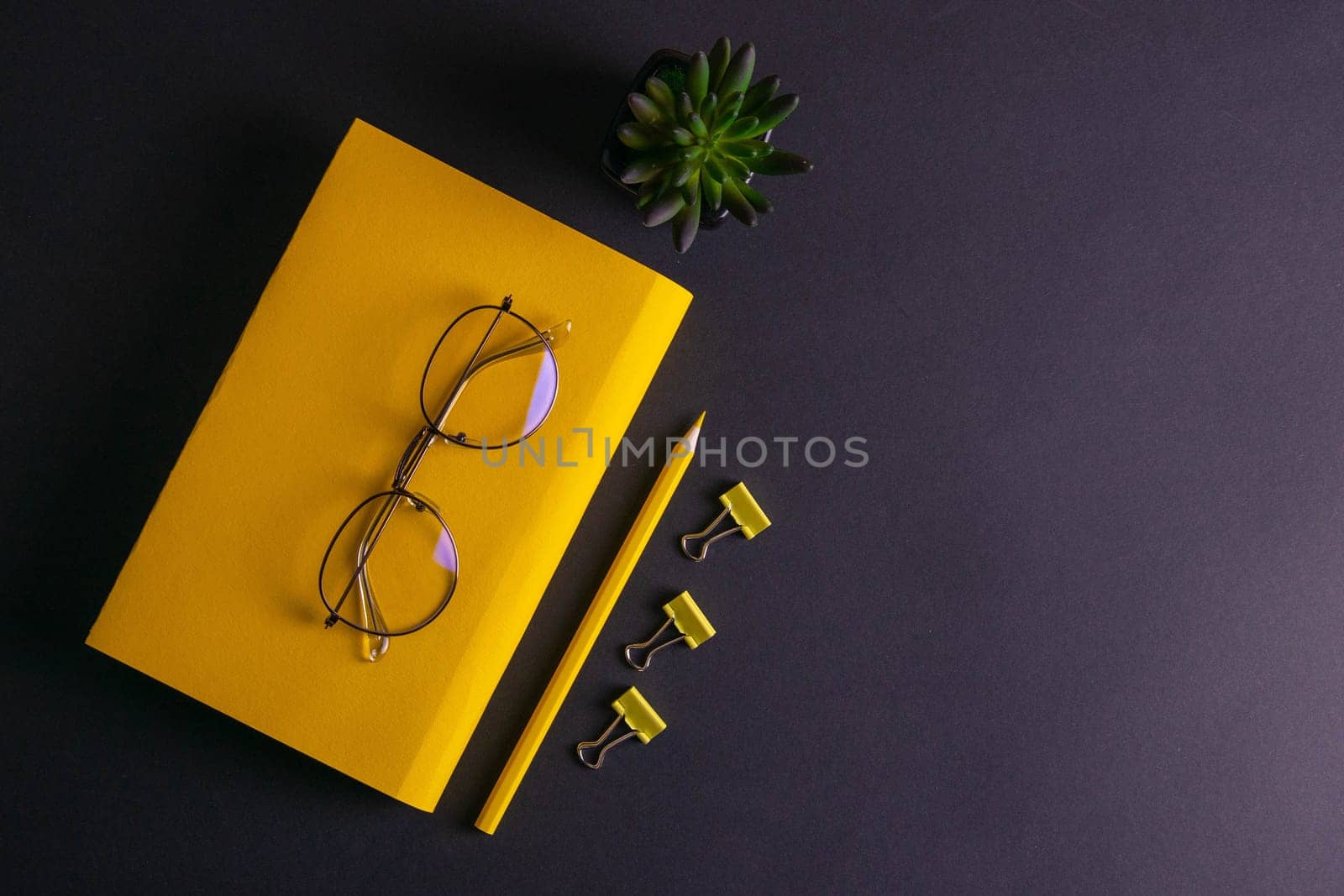 Yellow pencil, yellow notebook and glasses, pins, clip paper on on black background.