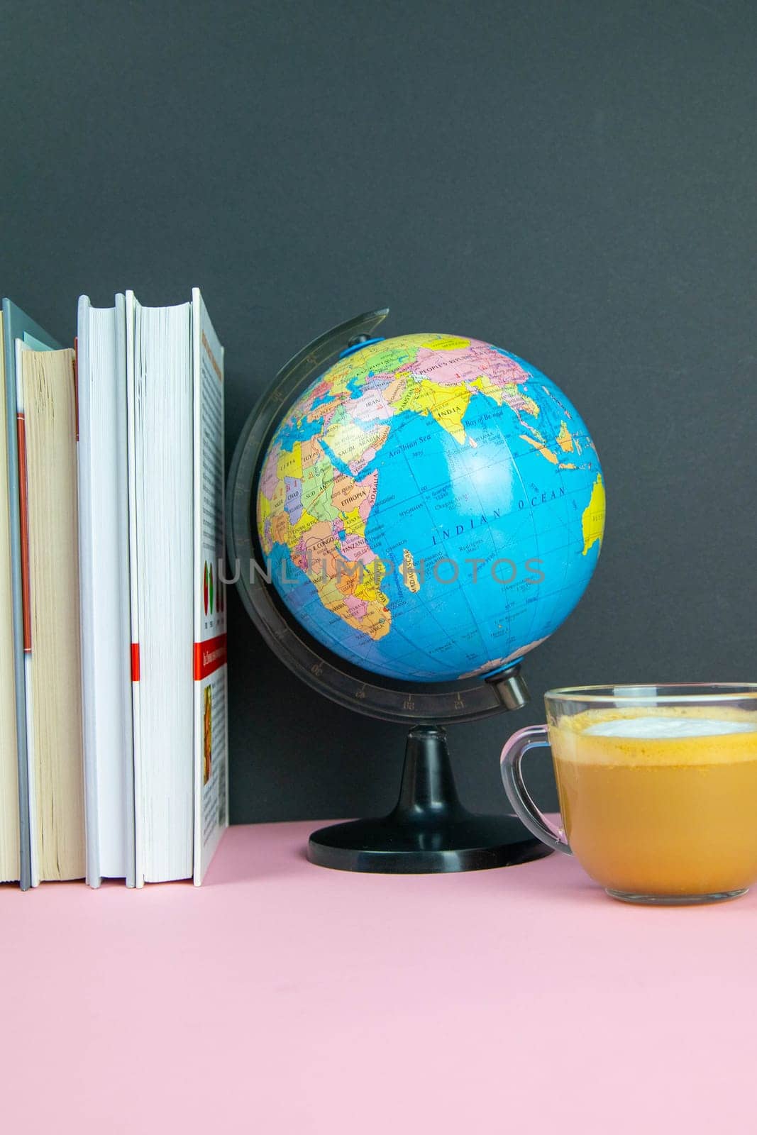 A stack of books, a globe and coffee with milk stand on a pink table.