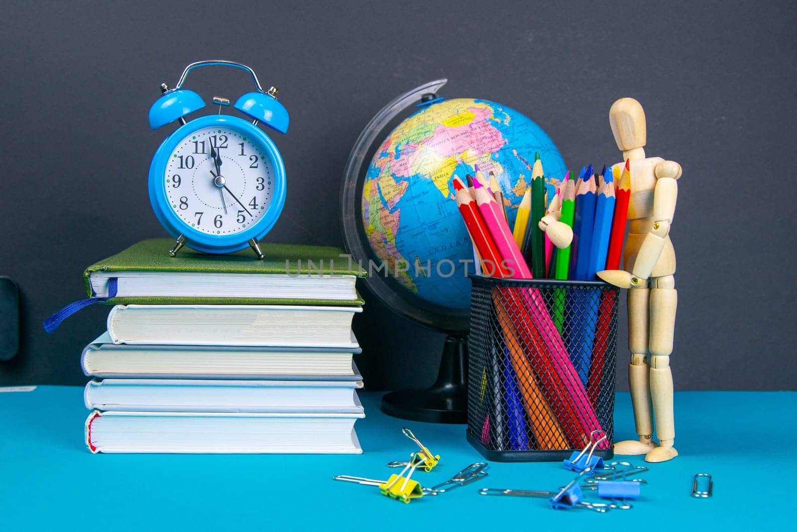 The globe and the clock stand on a stack of books, next to a glass of pencils and the pyramid is folded with wooden cubes and the figure of a wooden man hugs pencils.