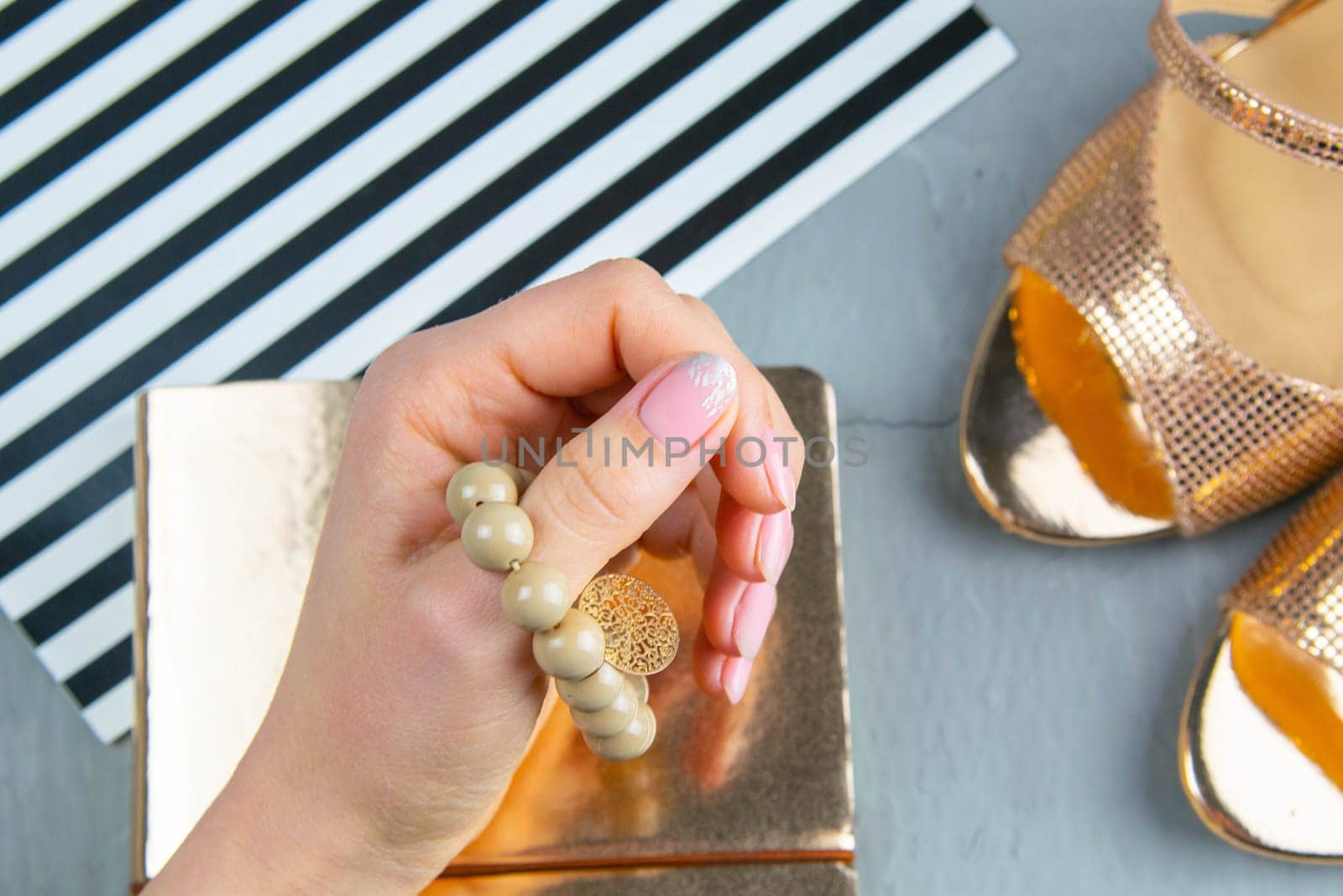 A woman's hand holds a bracelet on her finger over a gold notepad, next to them are gold shoes.