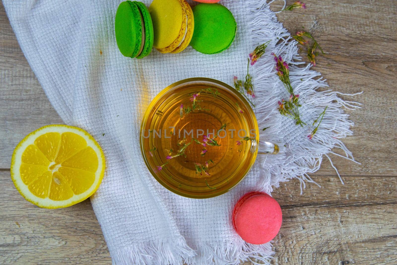 Glass transparent cup with mint tea stands on a wooden stand, next to a sugar bowl with sugar cubes, sliced lemon, spoon and multi-colored muffins.