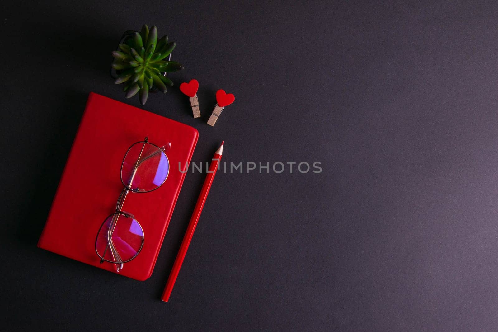 The red notebook stands on a black background, on the notebook there are glasses and next to the notebook, a flower, two clothespins in the shape of hearts and a red pencil.