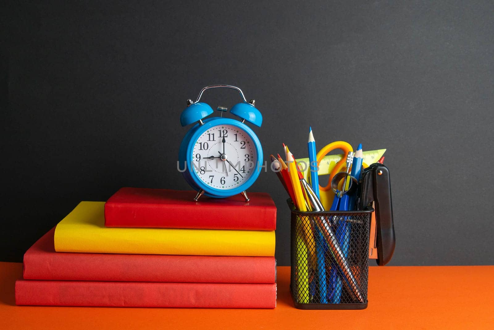 A stack of books, a green flower, blue clock and a glass of pencils stand on an orange background.