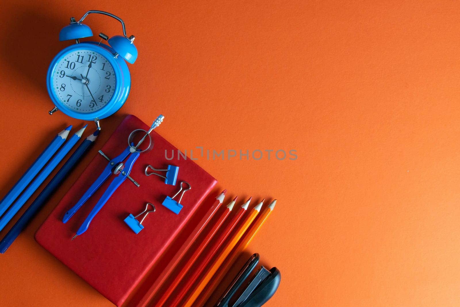 Red notebook with pencils, compass, stapler and clock on an orange background.