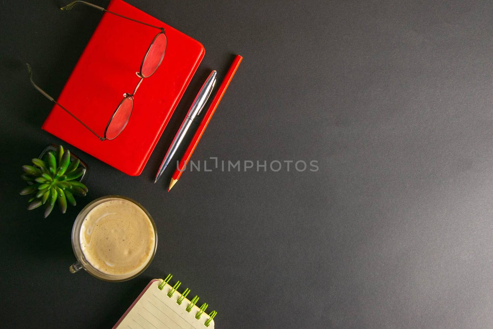 The red notebook stands on a black background, on the notebook there are glasses and next to the notebook, a flower, coffe, pen and a red pencil.