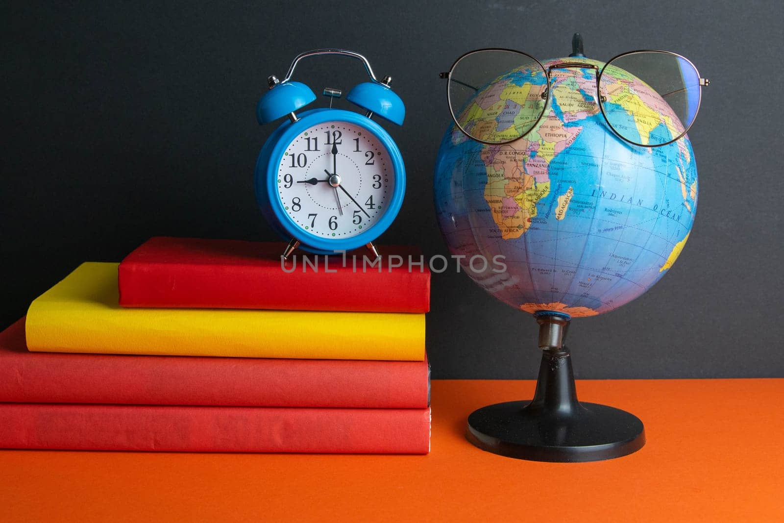 A stack of books, clock, world globe, and women's glasses in the foreground, stand on an orange background.
