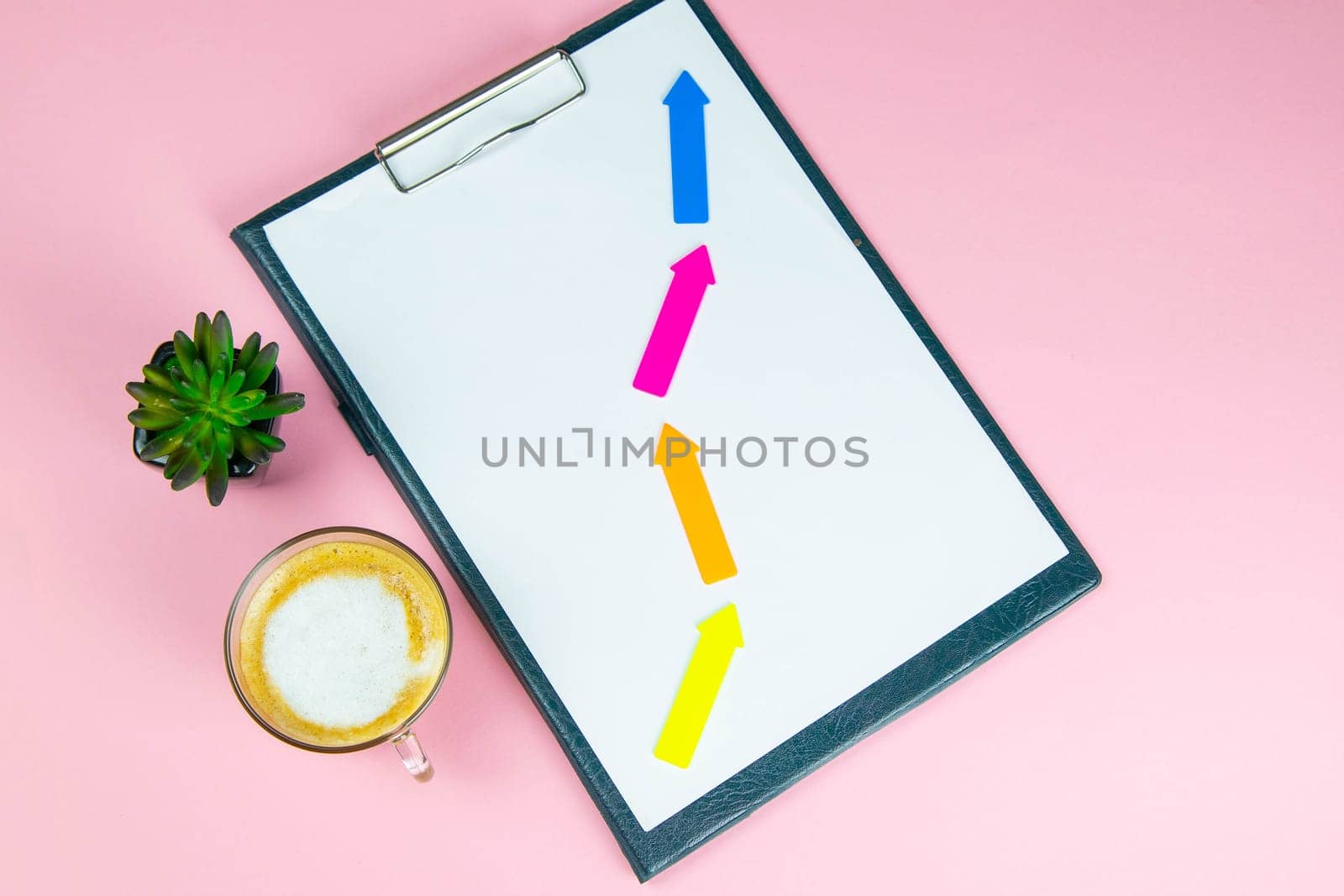 Tablet with a white sheet on which multicolored arrows show growth, success, next to stands a green plant and coffe with milk.