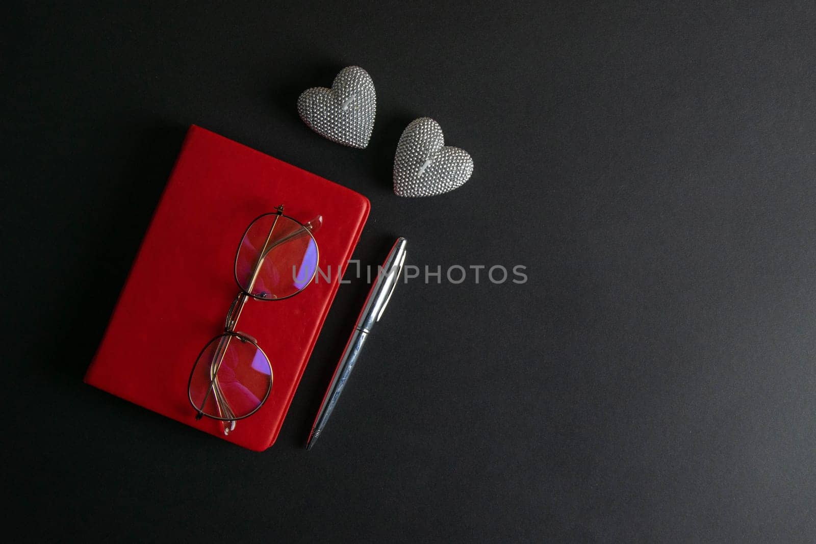 The glasses stand on a red notebook, there are two heart-shaped candles nearby, and a silver pen.