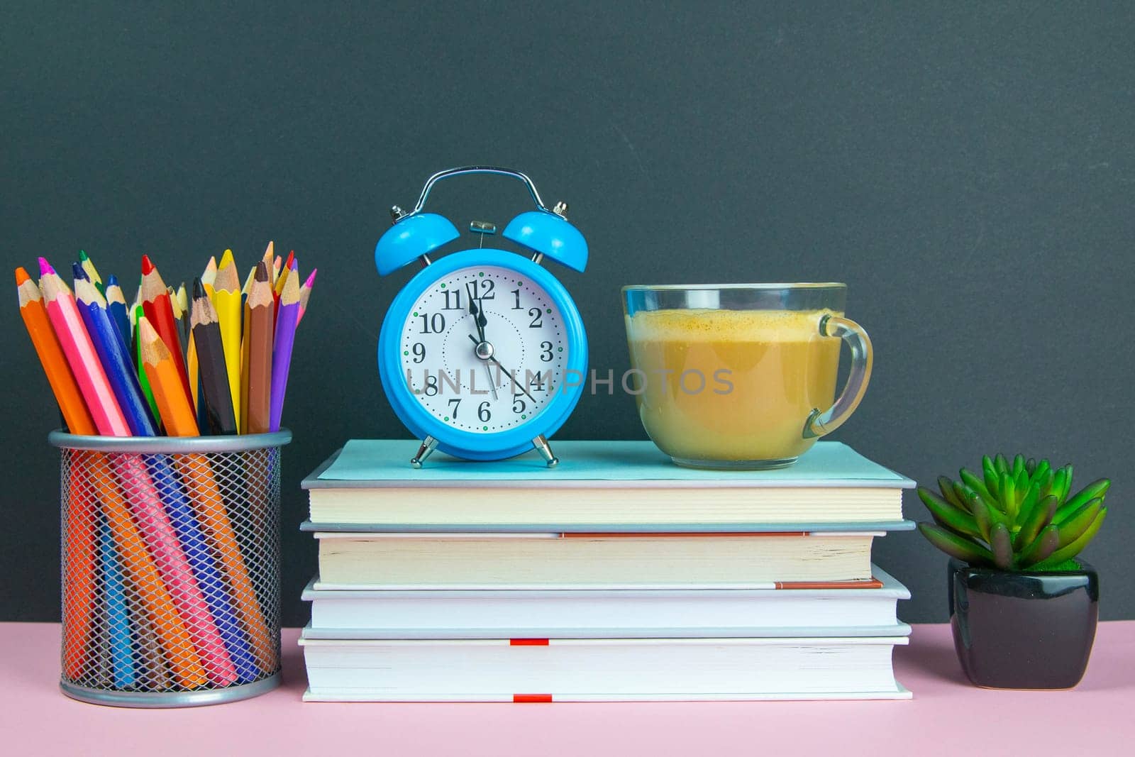 A stack of books on which stands a coffe with milk next to stands a green flower and a glass with pencils.