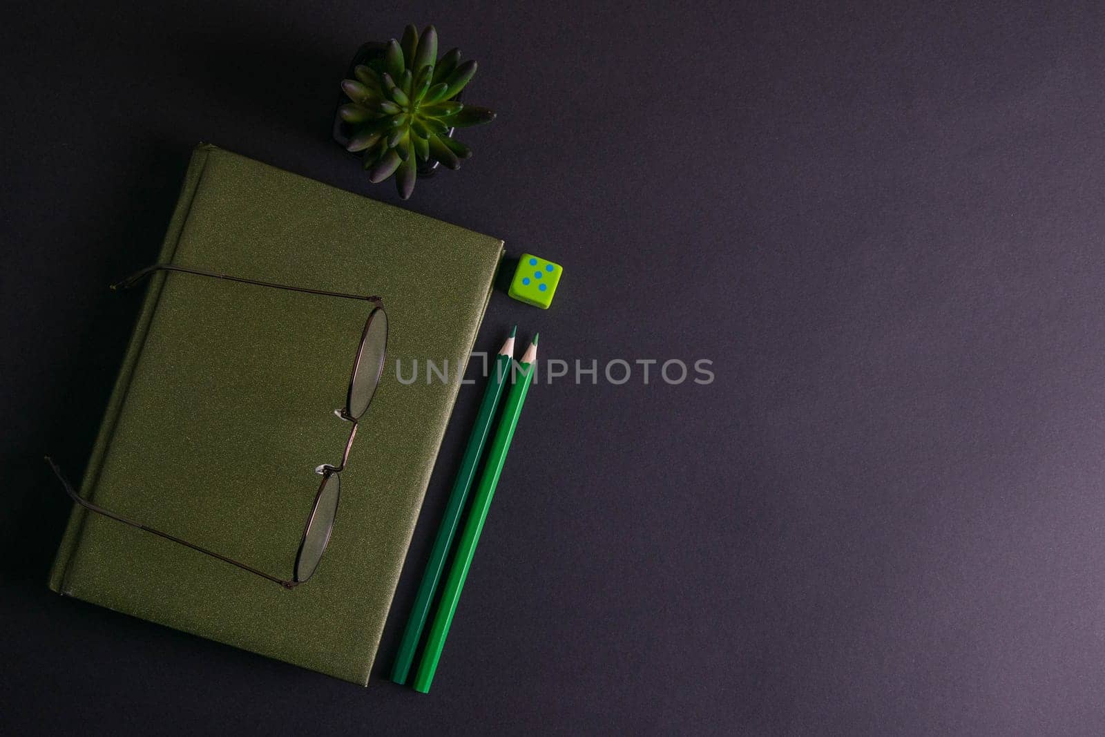 A green notebook with glasses on it has a green plant and green pencils next to the notebook.