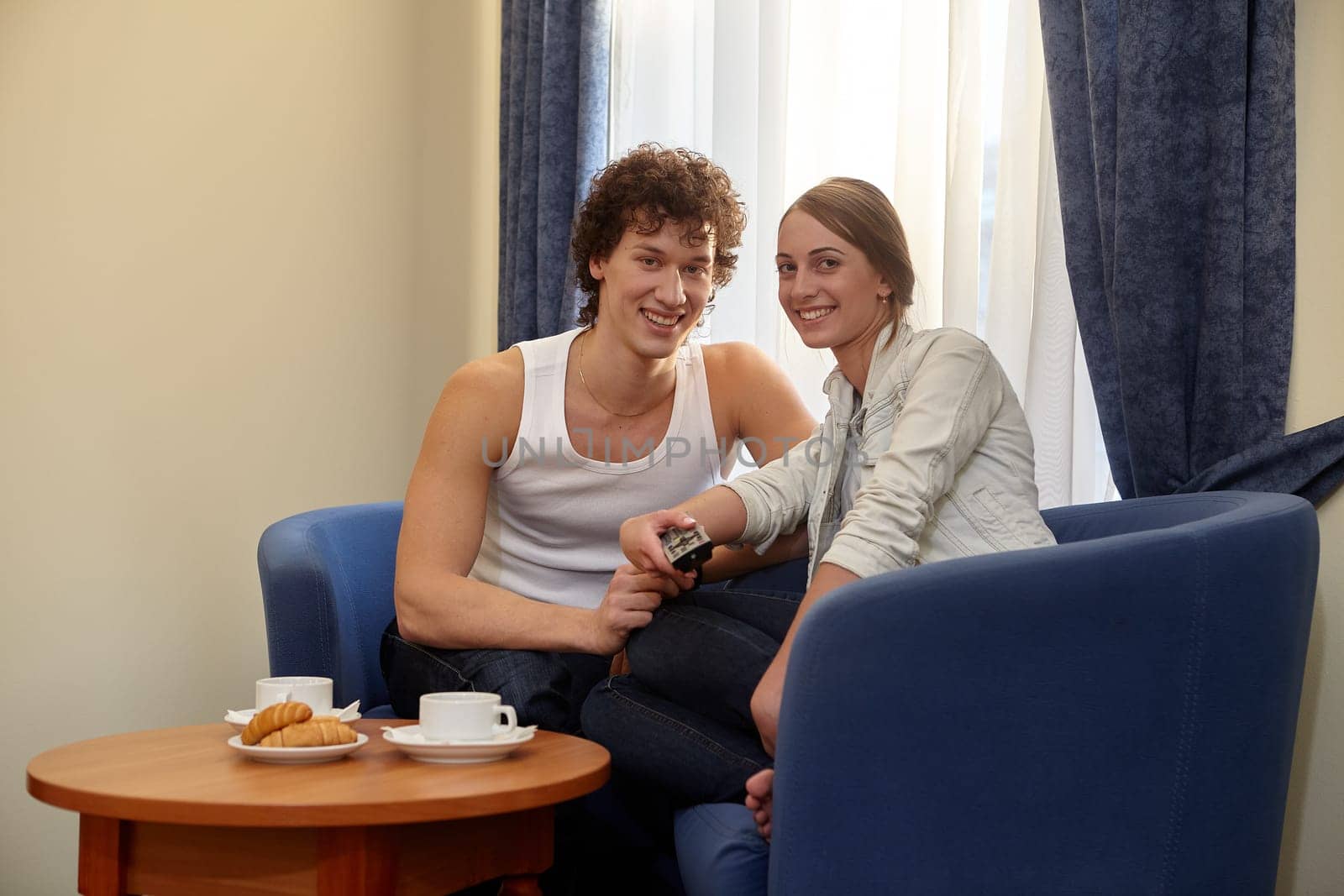 Happy young couple with breakfast are sitting in armchairs and watching TV in living room at home.