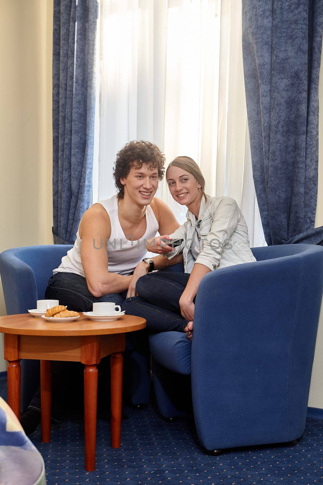 Happy young couple with breakfast are sitting in armchairs and watching TV in living room at home.