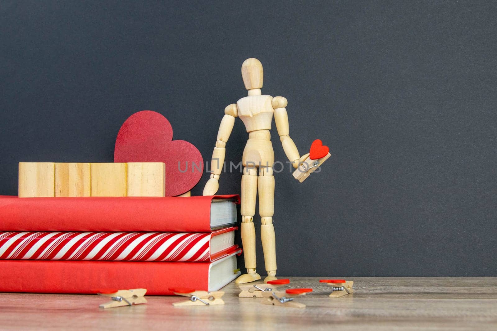 A stack of red books on which there are 4 wooden cubes, and next to there is a figure of a wooden man with a pin in the shape of a heart in his hand. Front views with clear space