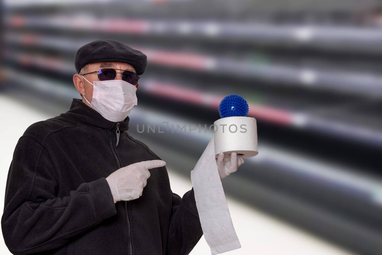 Coronavirus panic concept. Man with surgical mask, hood, glasses and medical gloves shows a roll of toilet paper with an abstract model of a virus covid-19 on it against blurred super market background.