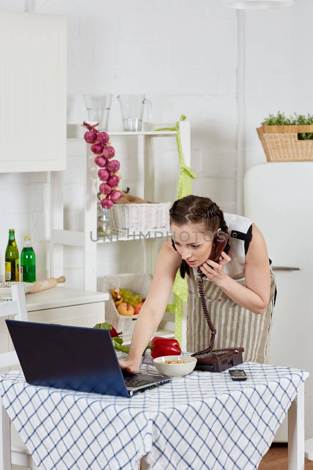 Woman in an apron with notebook and phone cooks in the kitchen. Isolation period, quarantine, social distancing. Remote education or remote work