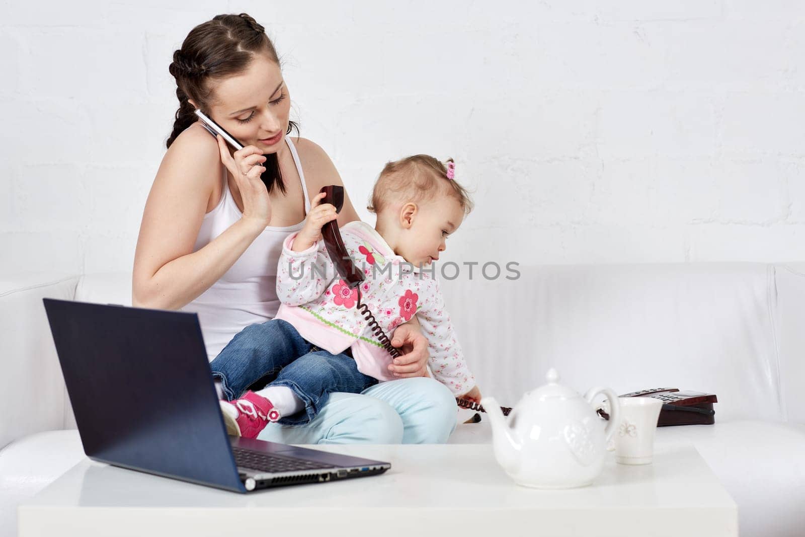 Small child and busy young woman with notebook and phone sit on the sofa in the room. Mother and baby. Isolation period, quarantine, social distancing. Remote work