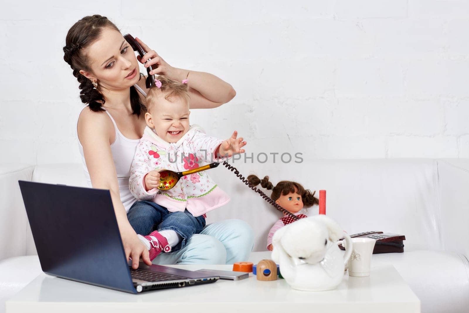 Small child and busy young woman with notebook and phone sit on the sofa in the room. Mother and baby. Isolation period, quarantine, social distancing. Remote work