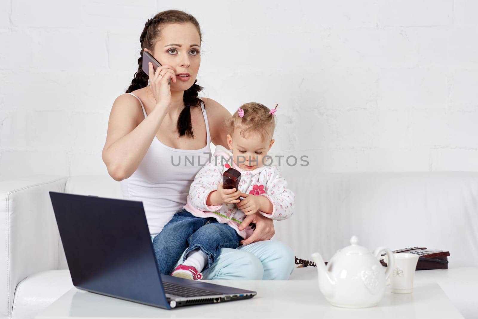Small child and busy young woman with notebook and phone sit on the sofa in the room. Mother and baby. Isolation period, quarantine, social distancing. Remote work