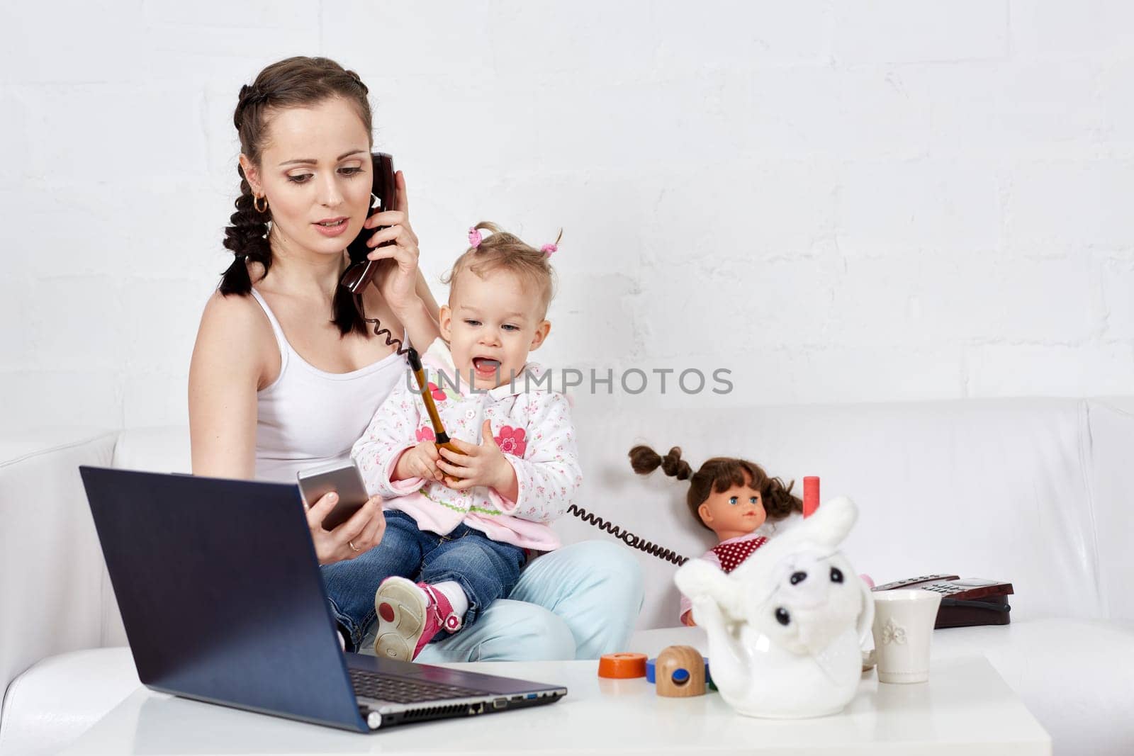 Small child and busy young woman with notebook and phone sit on the sofa in the room. Mother and baby. Isolation period, quarantine, social distancing. Remote work