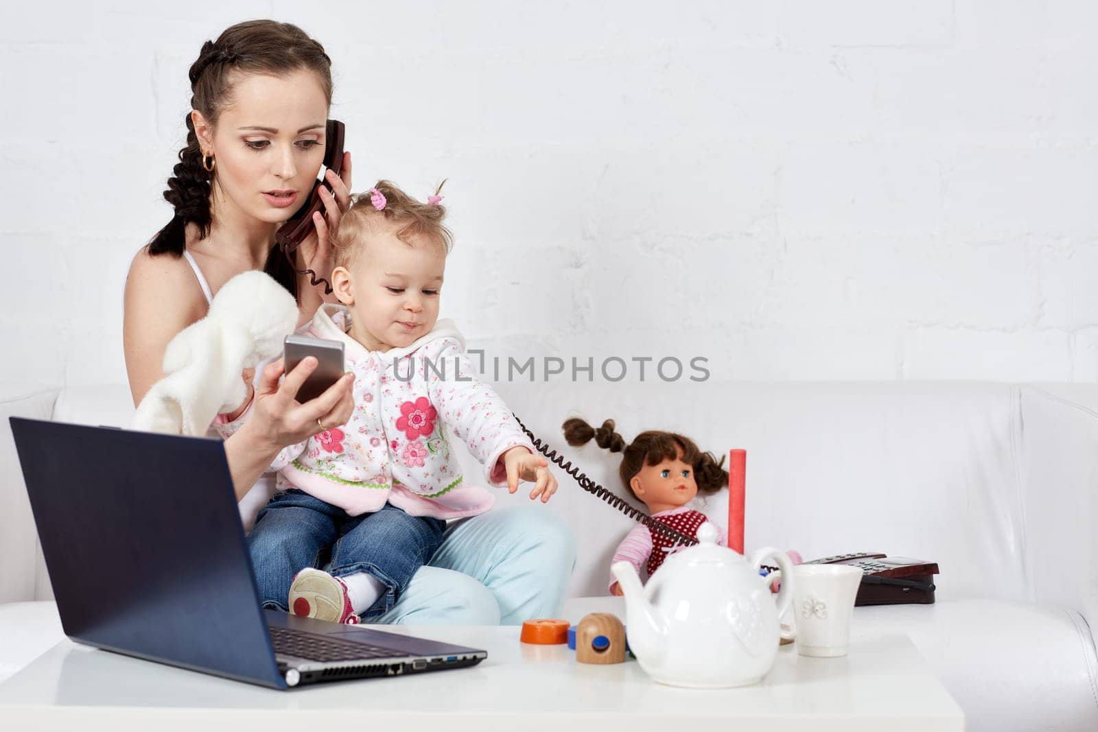 Small child and busy young woman with notebook and phone sit on the sofa in the room. Mother and baby. Isolation period, quarantine, social distancing. Remote work