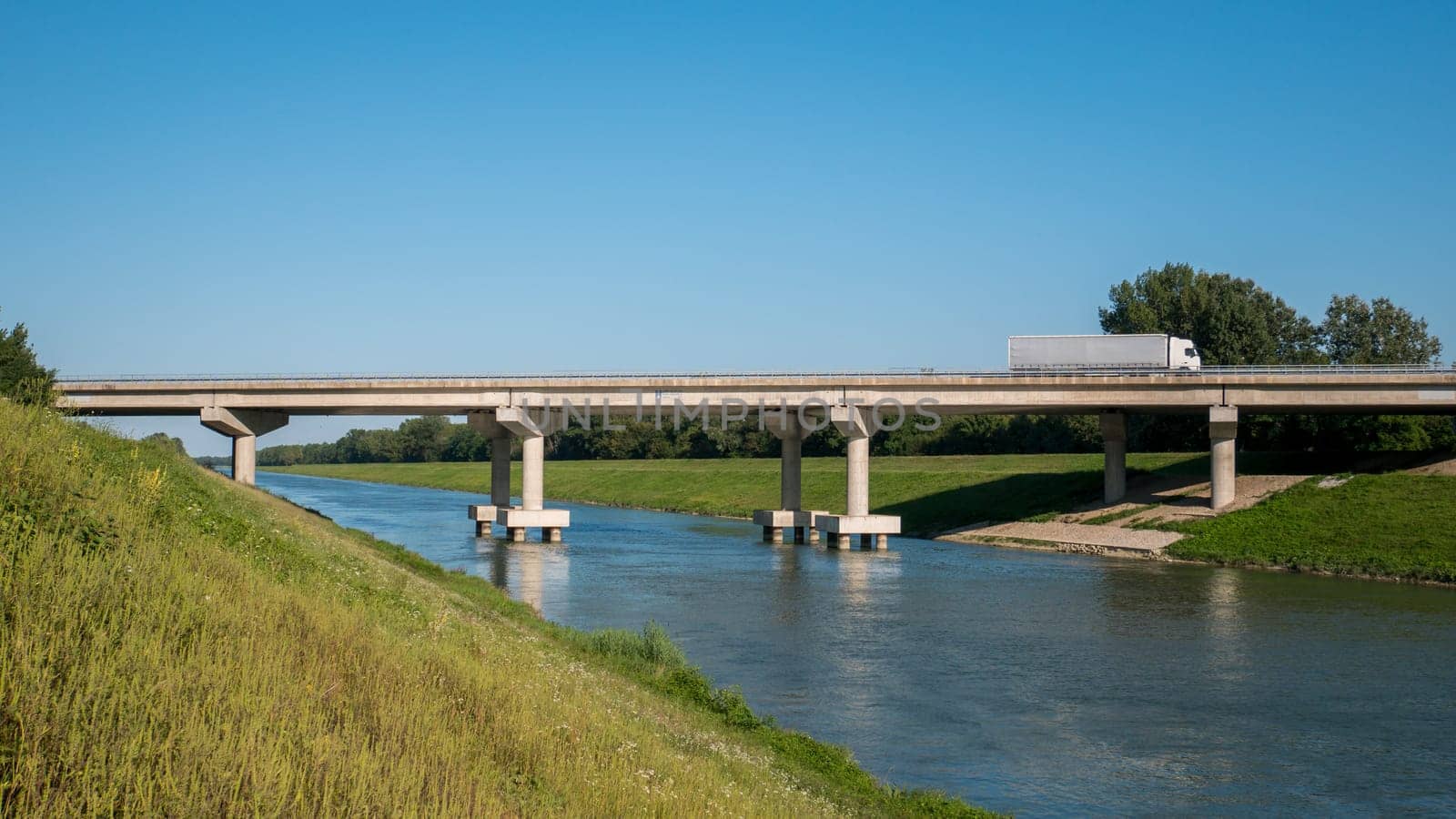 Bridge view and truck on the bridge. Pictured is a blue sky and a river, and a truck crossing the bridge over the river.