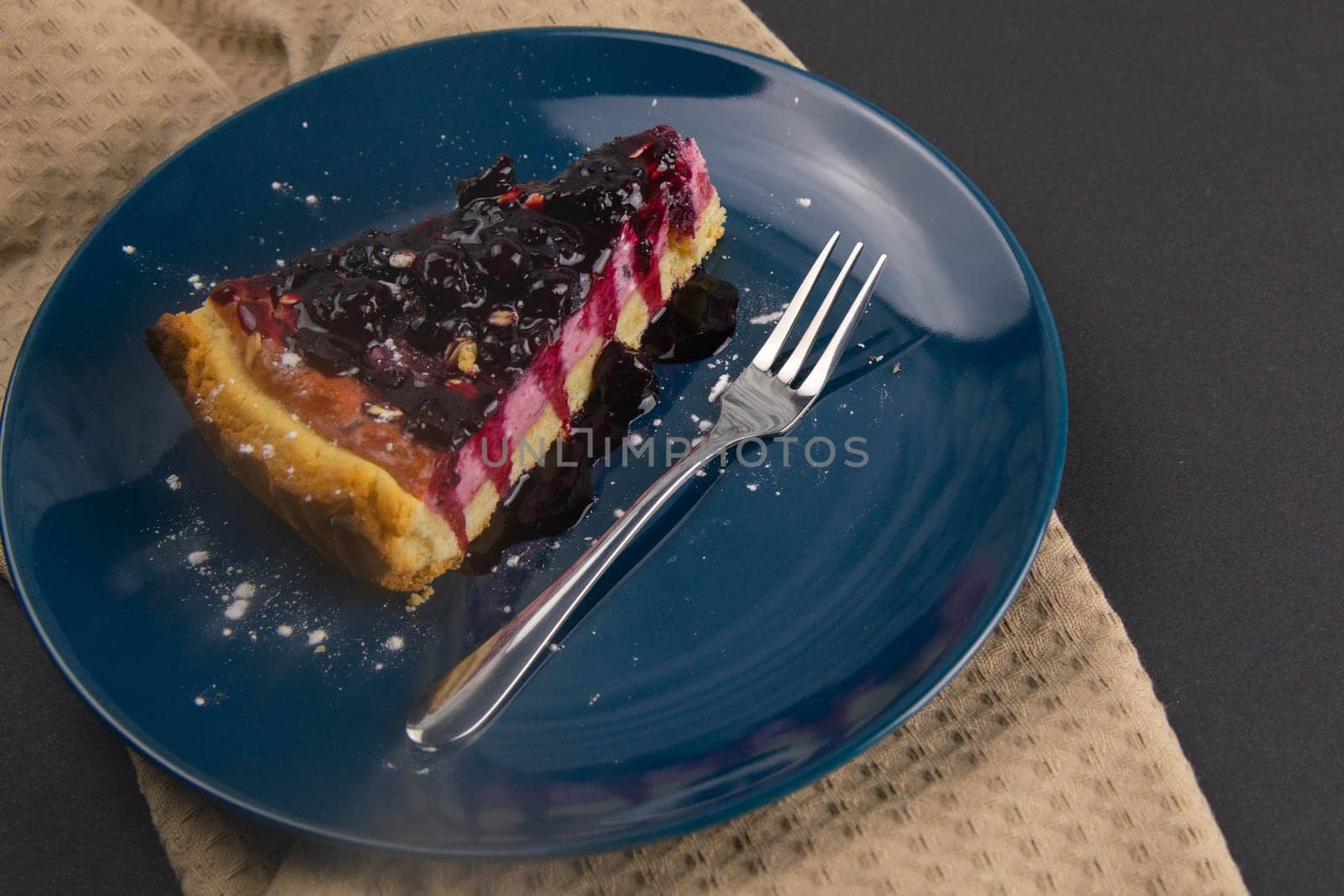 Slices of blueberry mousse cake with mirror glaze decorated berries and leaves of mint on a blue plate are on black table, close up. Top views.