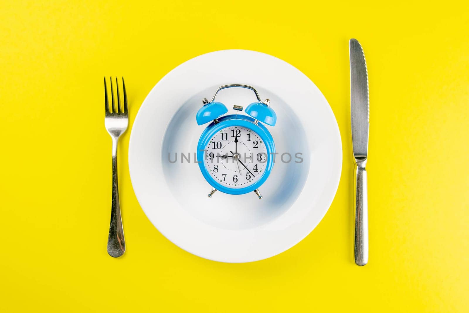 Alarm clock with fork and knife on the blue plate on blue table. Time to eat, Breakfast, Lunch Time and Dinner concept