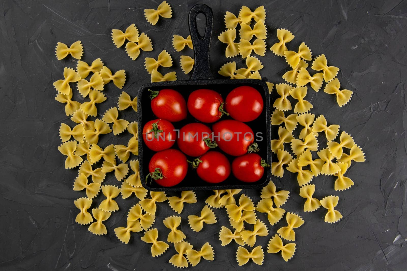 Raw pasta farfalle, tomatoes and frying pan on dark background. Top views, close-up.