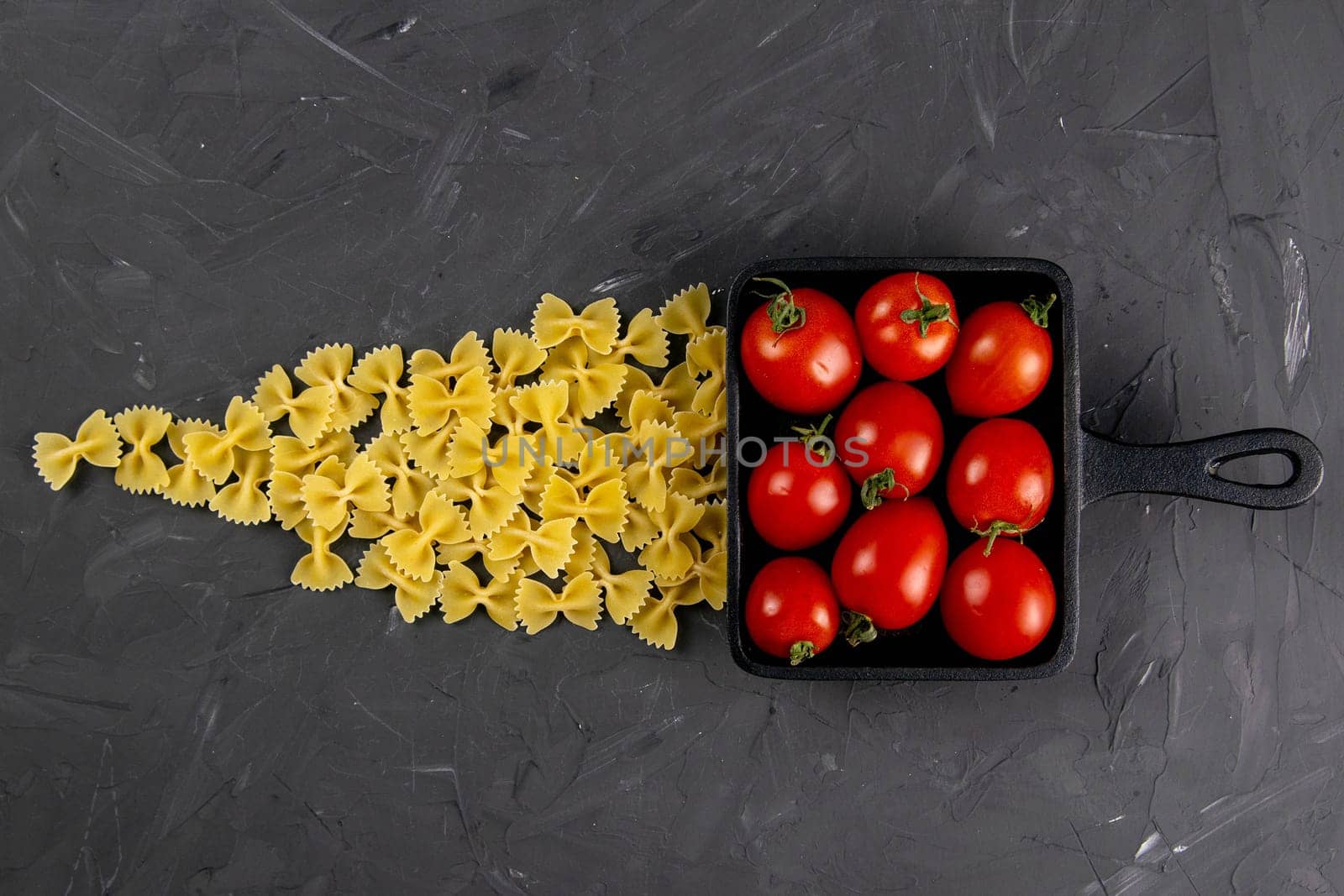 Raw pasta farfalle, tomatoes and frying pan on dark background. Top views, close-up.