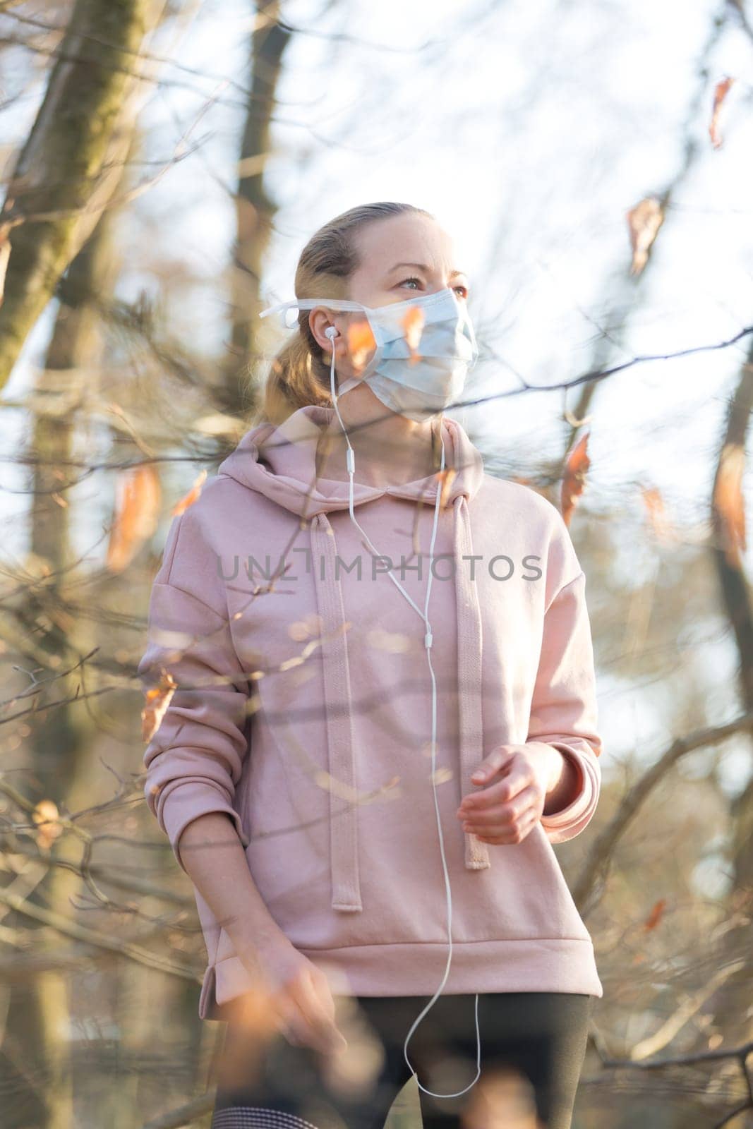 Portrait of caucasian sporty woman wearing medical protection face mask while walking in park, relaxing and listening to music. Corona virus, or Covid-19, is spreading all over the world.