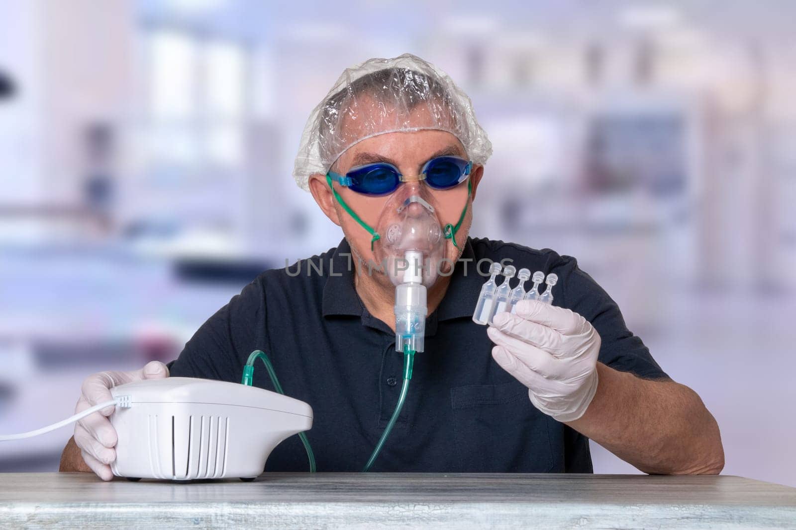 Coronavirus treatment concept. Man at table with nebulizer mask, safety goggles and medical hair hood and gloves looks at the ampoules over blurred hospital background. Protection of the respiratory tract.