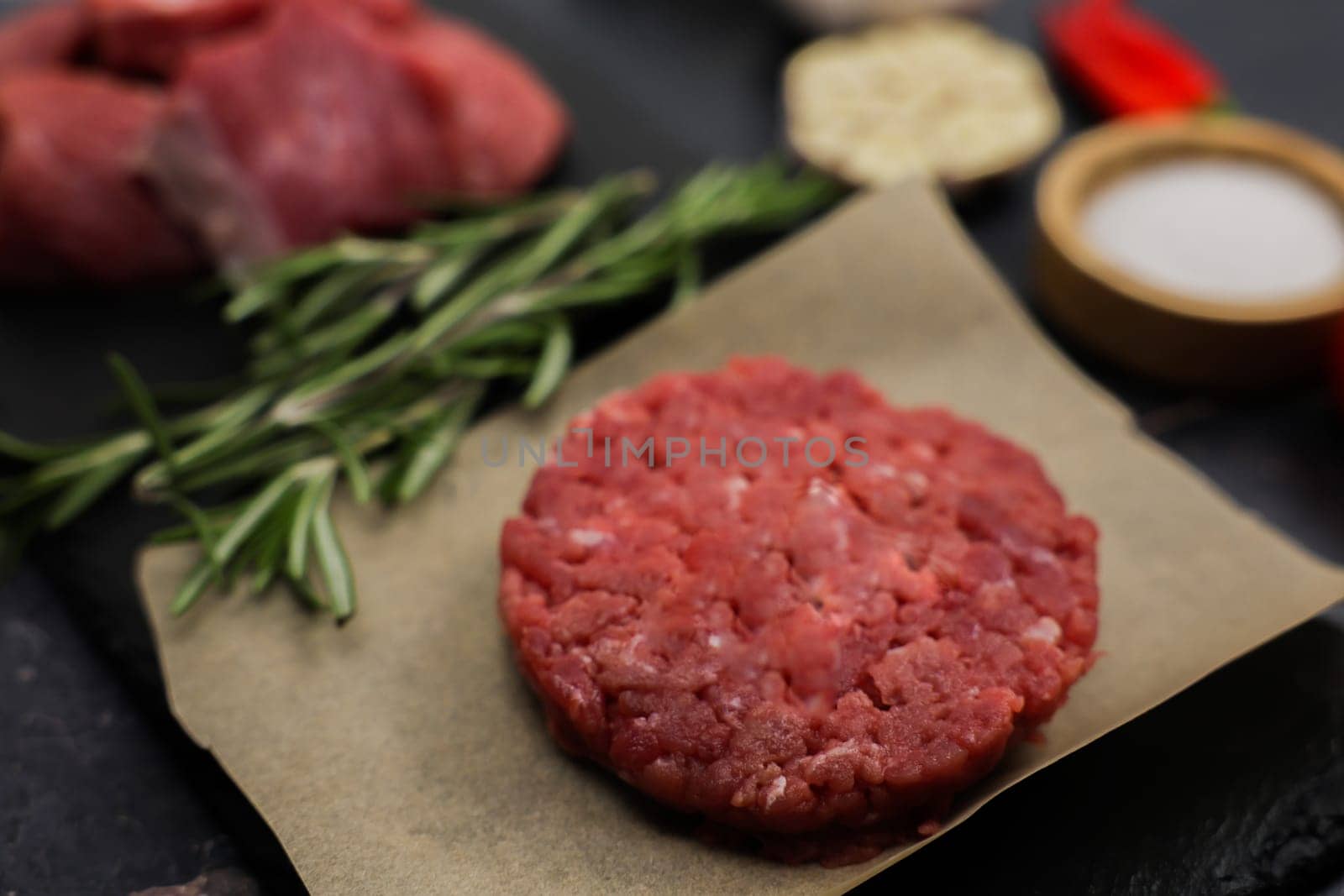 The concept of cooking a burger, fast food, natural products. Close-up beef raw round patty on paper surrounded by chunks of meat, seasoning, herbs and vegetables out of focus on dark background. High quality photo