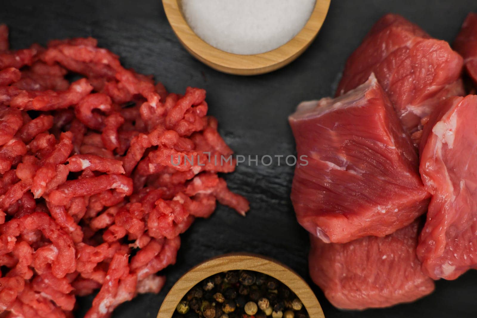 A close-up view from above of chunks of beef meat and red minced meat with streaks of fat lying on a black background with spices and salt in wooden bowls on the sides. The concept of cooking, shredding meat, diet, natural products. High quality photo