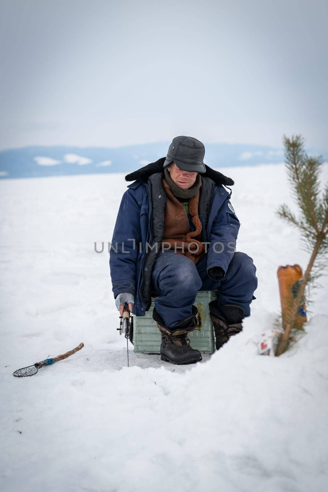 Baikal lake, Siberia, Russia - March 05, 2020 : Winter ice fishing. A fisherman is fishing on spinings near tents and cars on Lake Baikal on a March day. Baikal lake, Siberia, Russia