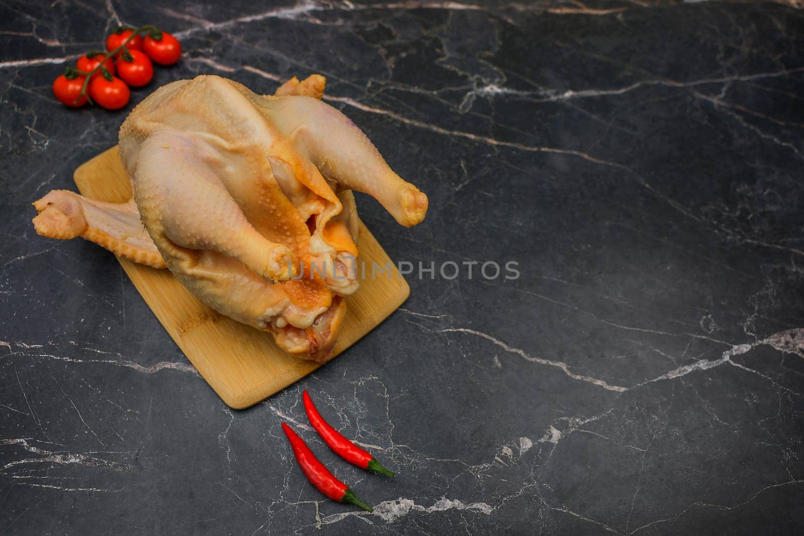 A chicken carcass, butchered and plucked, lies on a wooden board against a background of black marble, surrounded by cherry tomatoes and hot red peppers with space for text. The concept of cooking, poultry meat, dietary dishes. High quality photo