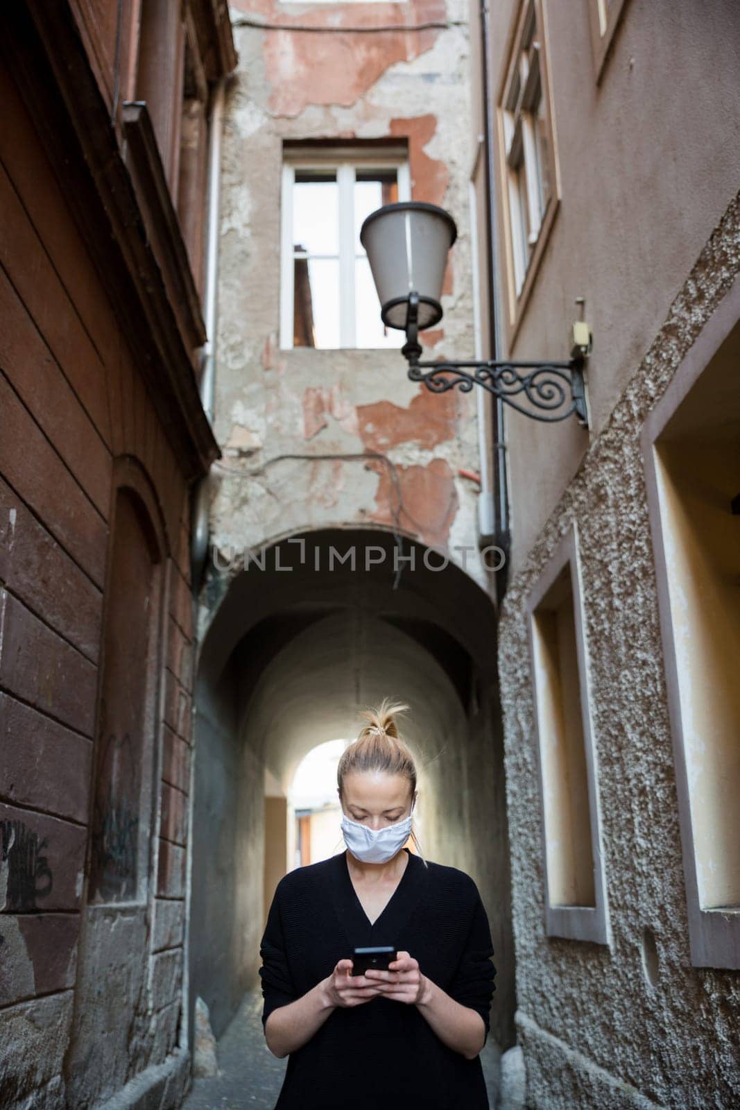 COVID-19 pandemic coronavirus. Casual caucasian woman at medieval city street using mobile phone, wearing protective face mask against spreading of coronavirus and disease transmission.