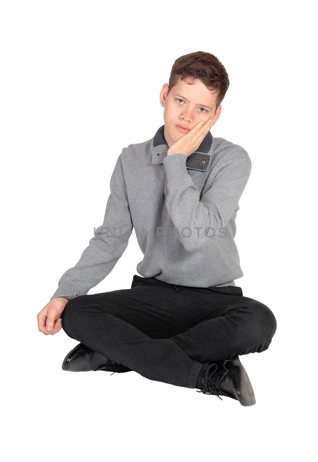 A handsome young teenage boy sitting in a gray sweater on the floor .in the studio with one hand on his face, isolated for white background.
