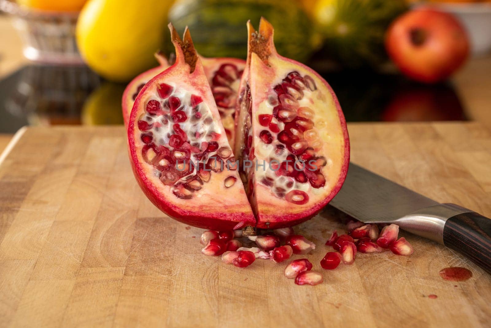 Close up of two half pomegranates fruits and a damascus kitchen knife on a wooden cutting board with pomegranate seeds in the foreground in a domestic kitchen