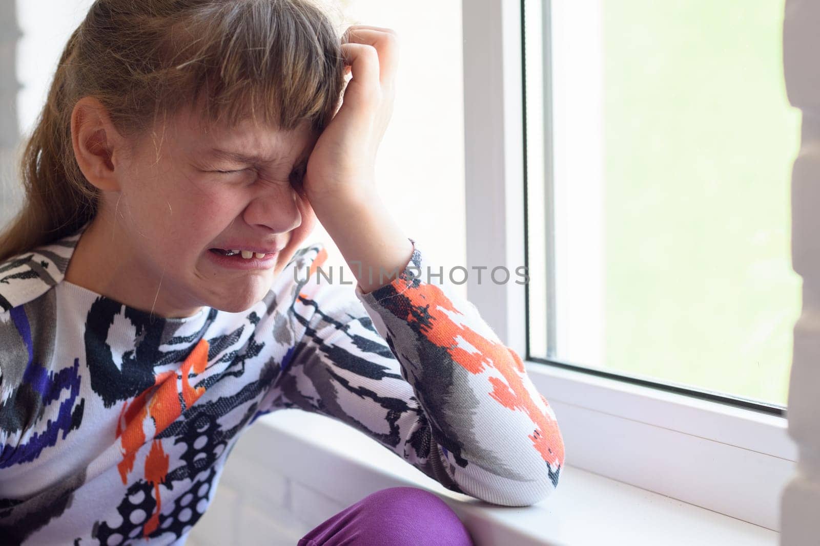 Teen girl crying a lot while sitting by the window in the room, close-up