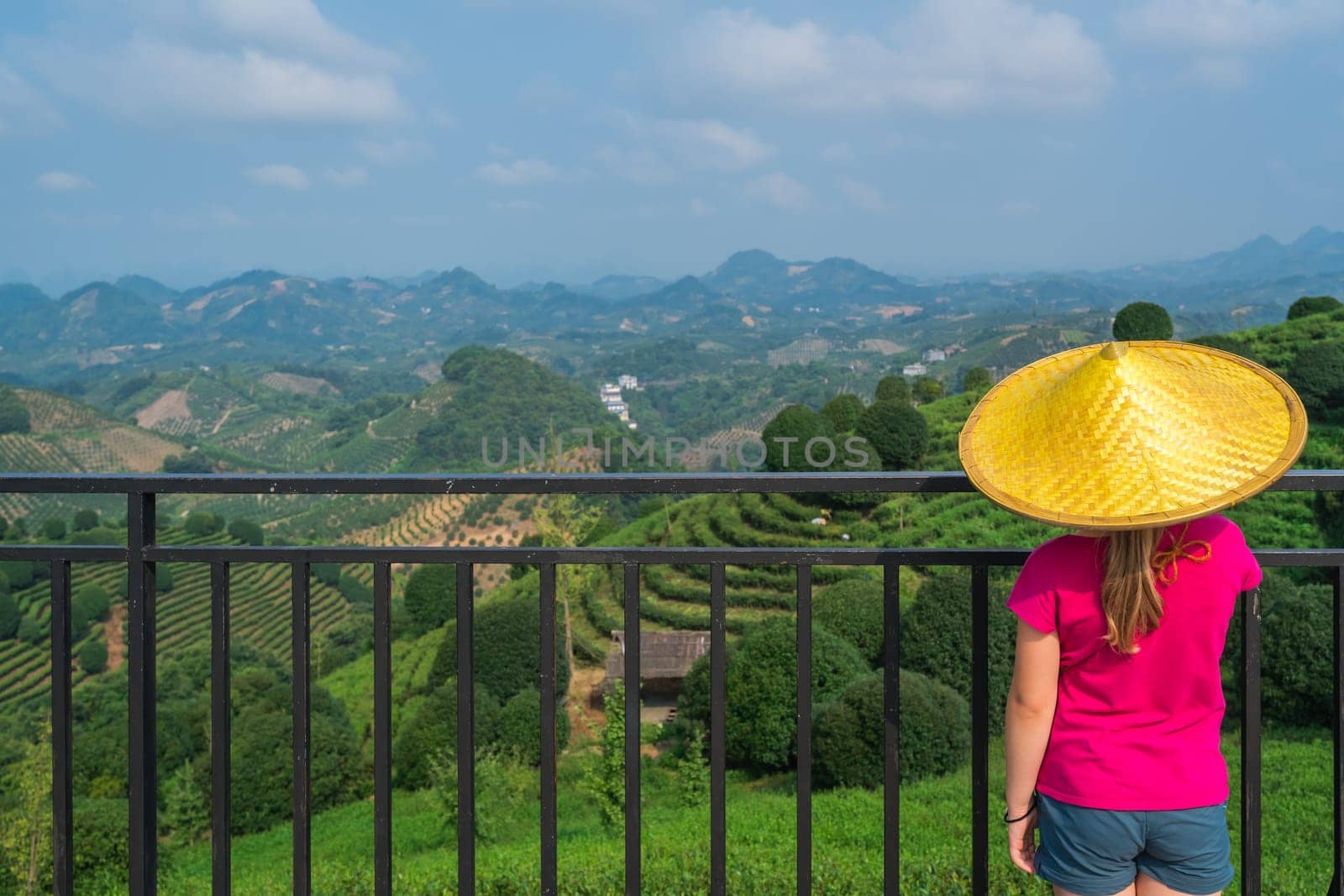 A beautiful closeup of a female leaning on the fences in Asian conical hat against a foliage forest and mountains