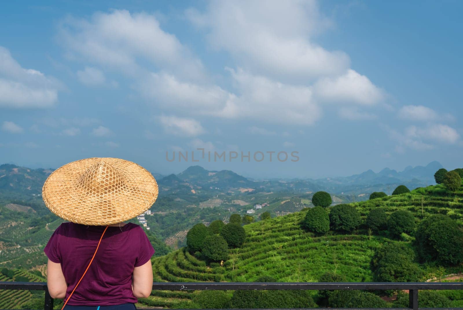 A beautiful closeup of a female leaning on the fences in Asian conical hat against a foliage forest, mountains and terraced tea plantation