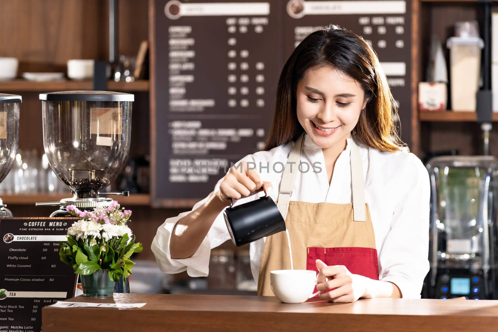 Young adult asian female woman barista pouring fresh milk to prepare latte coffee for customer in cafe bar with her colleague working in background. For small business startup in food industry concept