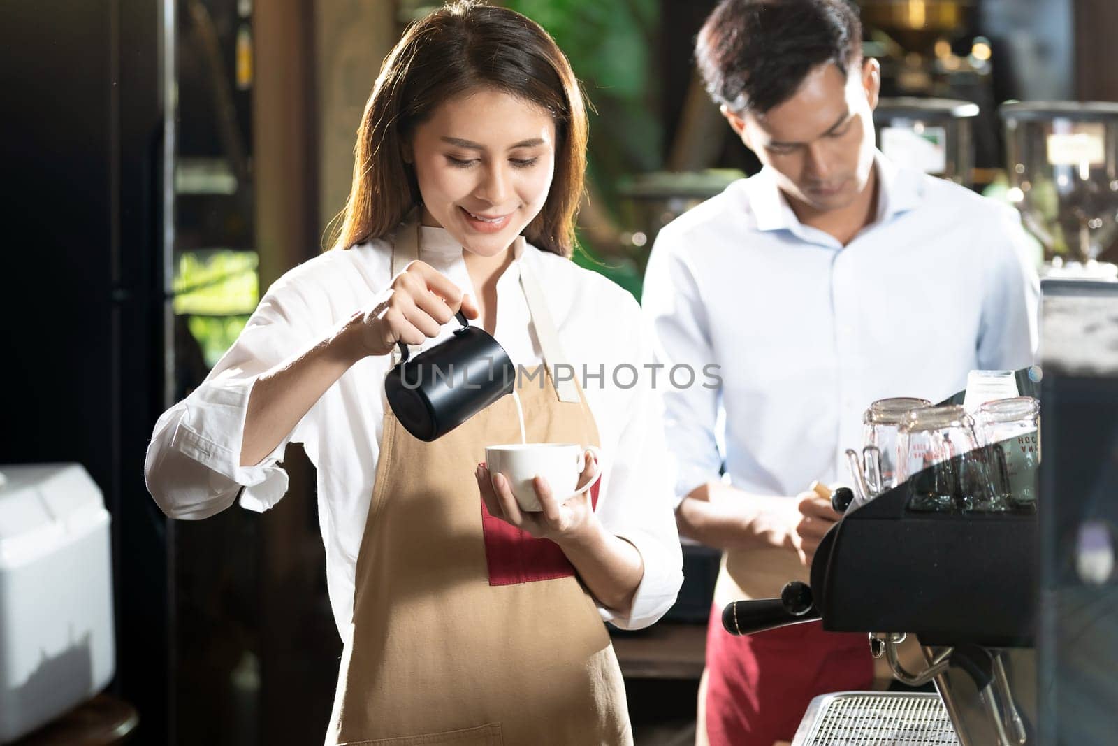 Young adult asian female woman barista pouring fresh milk to prepare latte coffee for customer in cafe bar with her colleague working in background. For small business startup in food industry concept