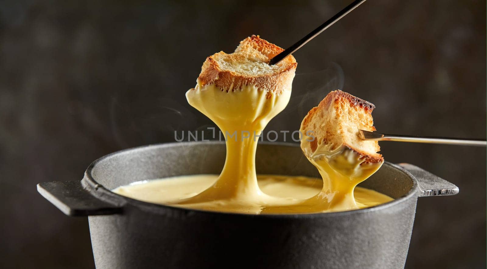 Delicious melted cheese fondue in winter served in a cast iron pot with forks dipping toasted baguette and rising steam over a dark background
