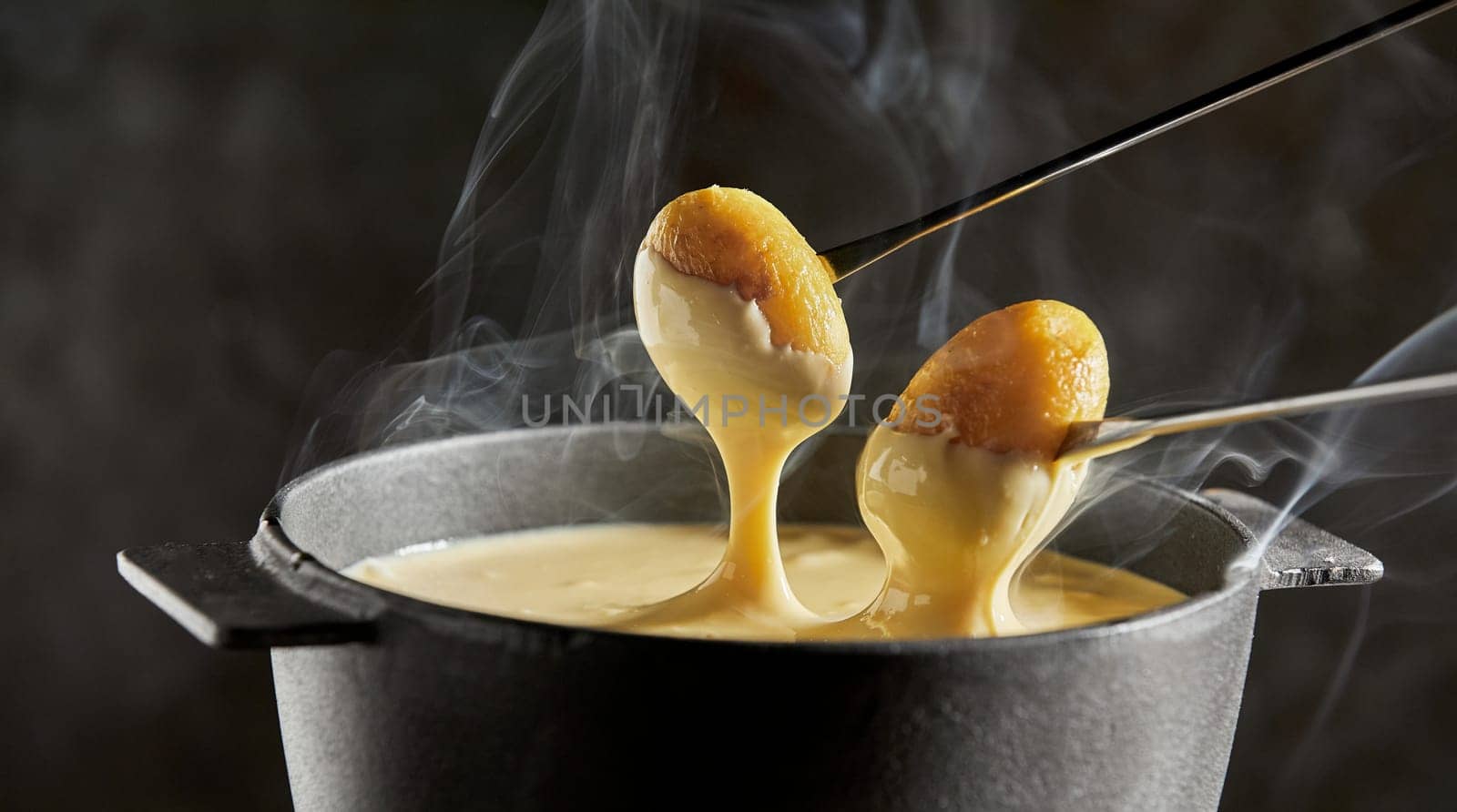 Dipping baby potatoes into a steaming cheese fondue in a cast iron pot in close up over a dark background
