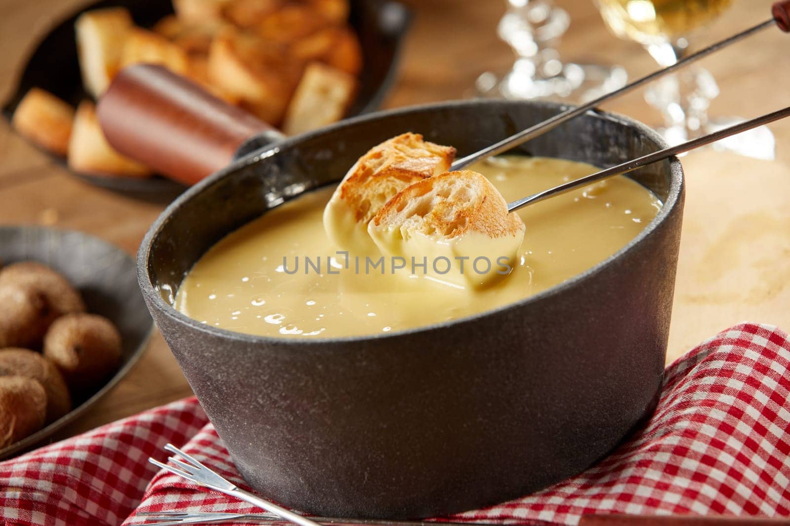 Dipping toasted baguette into a hot cheese fondue served in a cast iron pot on a rustic table with accompaniments