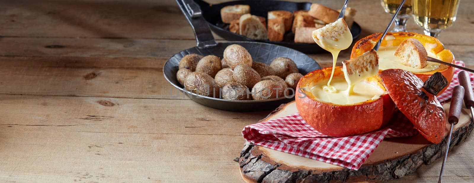Enjoying an autumn Swiss cheese fondue with glasses of wine and accompanying toast and potatoes served in a hollow pumpkin on a rustic table in a panorama banner