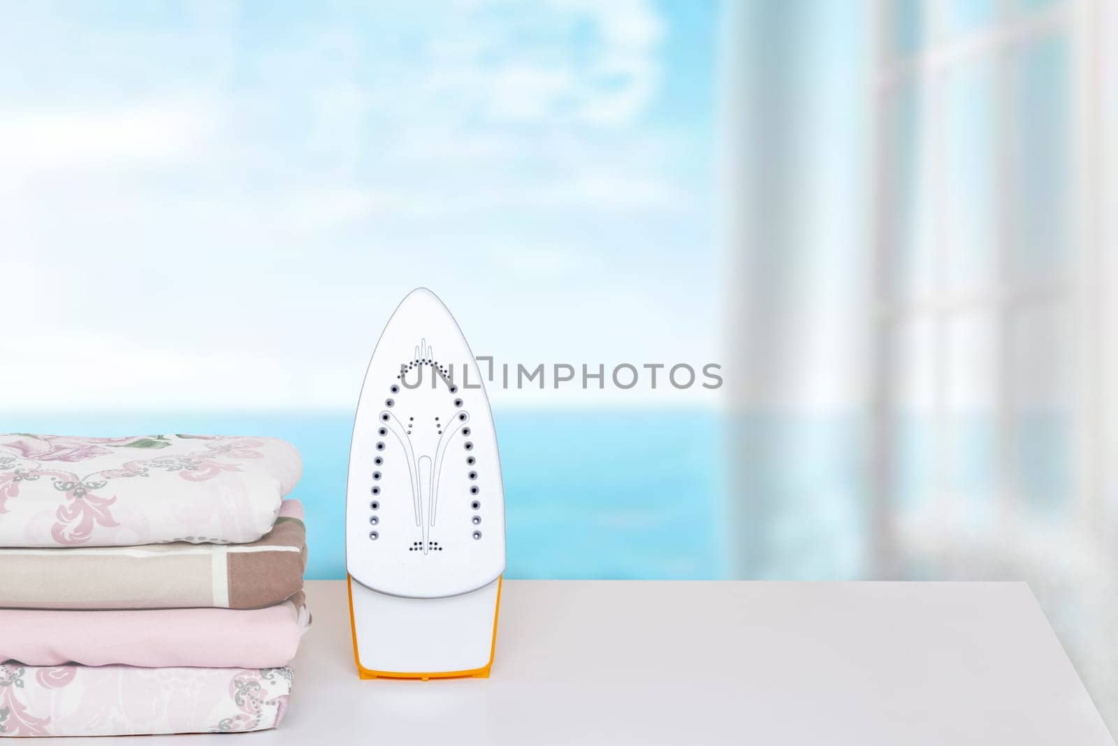 Household laundry ironing. Close-up of a yellow electrical iron and a stack of ironed clothes on an ironing board indoors over abstract blurred natural ocean background. Advertising for the laundry service with space.